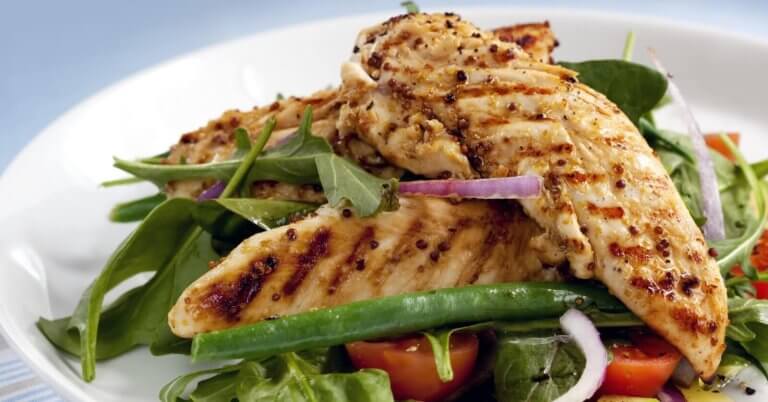 A plate of grilled chicken breasts served on a bed of fresh mixed greens, including arugula and green beans, is now available through healthy food delivery in Manila. The dish is garnished with slices of red onion and halves of cherry tomatoes. The chicken has a seared, seasoned exterior, and the salad appears crisp and vibrant.