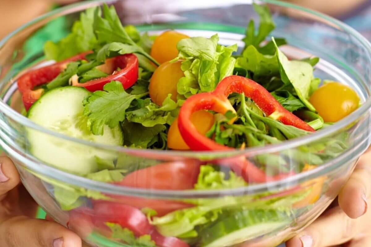 A person is holding a clear glass bowl filled with a fresh salad. The salad includes slices of red bell pepper, rounds of cucumber, yellow cherry tomatoes, arugula, and mixed greens. Another person wearing a blue and green striped shirt is partially visible in the background at one of Metro Manila's healthy restaurants.