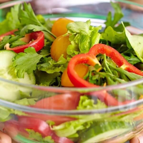 A person is holding a clear glass bowl filled with a fresh salad. The salad includes slices of red bell pepper, rounds of cucumber, yellow cherry tomatoes, arugula, and mixed greens. Another person wearing a blue and green striped shirt is partially visible in the background at one of Metro Manila's healthy restaurants.