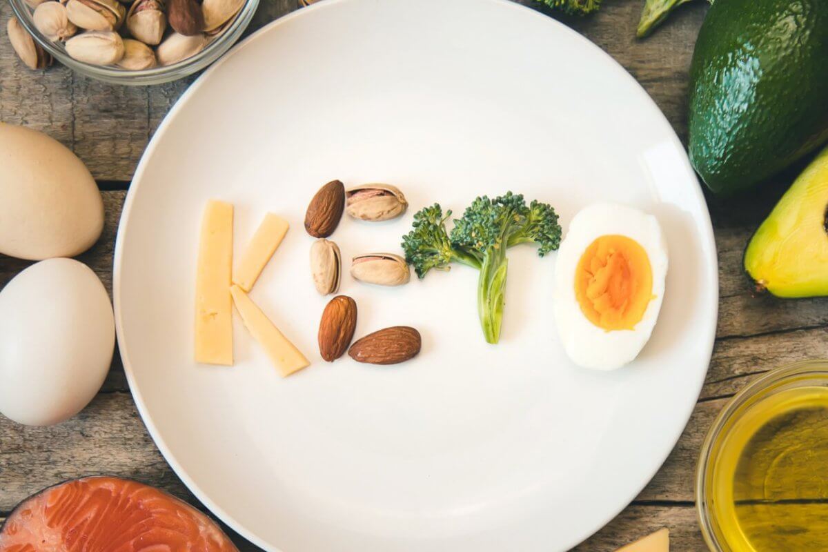 A white plate on a wooden table spells out "Keto" using various foods: cheese for "K", almonds and pistachios for "e", broccoli for "t", and a deviled egg for "o". Surrounding the plate are keto-friendly ingredients like eggs, an avocado, salmon, nuts, broccoli, and a bowl of oil. Discover fresh keto supplies in Metro Manila shops.