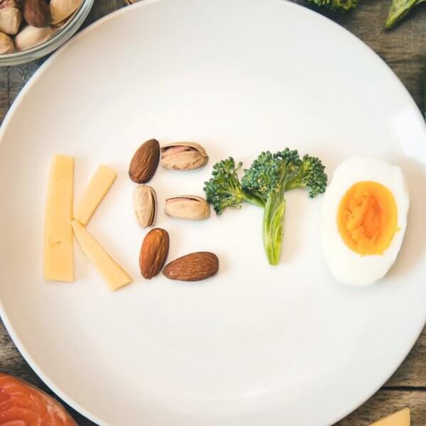 A white plate on a wooden table spells out "Keto" using various foods: cheese for "K", almonds and pistachios for "e", broccoli for "t", and a deviled egg for "o". Surrounding the plate are keto-friendly ingredients like eggs, an avocado, salmon, nuts, broccoli, and a bowl of oil. Discover fresh keto supplies in Metro Manila shops.