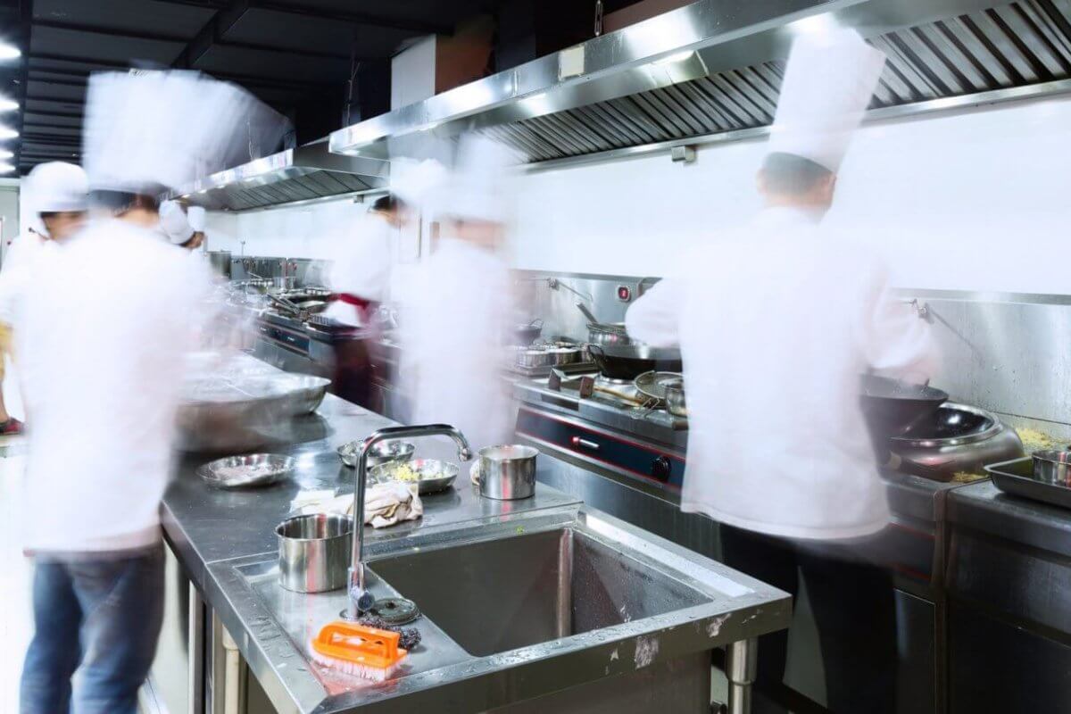 A bustling commercial kitchen with stainless steel counters, sinks, and ventilation hoods. Several chefs in white uniforms and hats are blurred, indicating rapid movement as they address every culinary need. Pots, pans, and utensils from Top 50 Kitchen Supply Stores in Metro Manila are scattered across the workspaces.