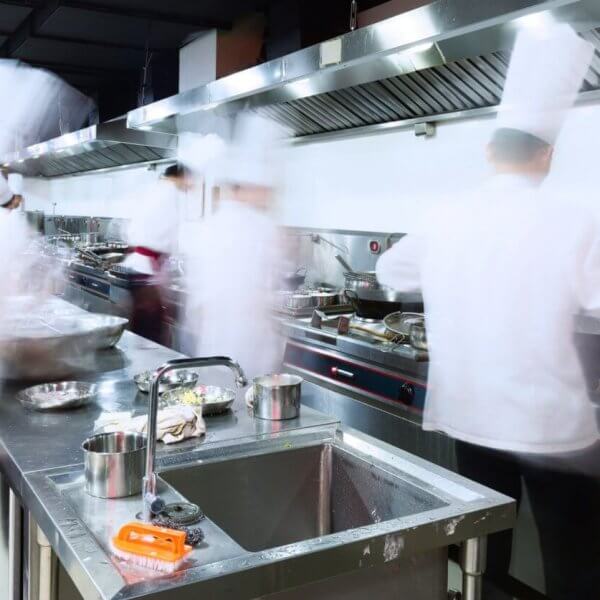 A bustling commercial kitchen with stainless steel counters, sinks, and ventilation hoods. Several chefs in white uniforms and hats are blurred, indicating rapid movement as they address every culinary need. Pots, pans, and utensils from Top 50 Kitchen Supply Stores in Metro Manila are scattered across the workspaces.