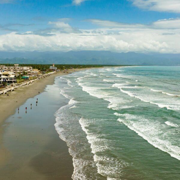 An aerial view of a long, sandy beach on Luzon with gentle waves rolling in from a turquoise ocean. On the left side, buildings and palm trees that are popular tourist attractions line the shore, while a few people are scattered along the wet sand. In the background, lush mountains are partially obscured by clouds under a bright, blue sky.