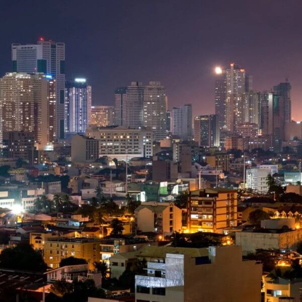 Makati cityscape at dusk featuring numerous high-rise buildings illuminated by varied lights defines the bustling spirit of Makati. The foreground consists of lower residential buildings with streetlights glowing, near Barangay Halls. The sky transitions from purple to blue, reflecting off some skyscrapers.