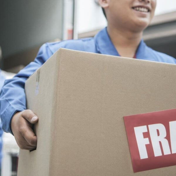 A person smiling and holding a large cardboard box with a red "FRAGILE" label. In the background, another person in a similar uniform is unloading a box from a truck. The setting appears to be one of the best moving companies in Metro Manila, providing top-notch delivery service.