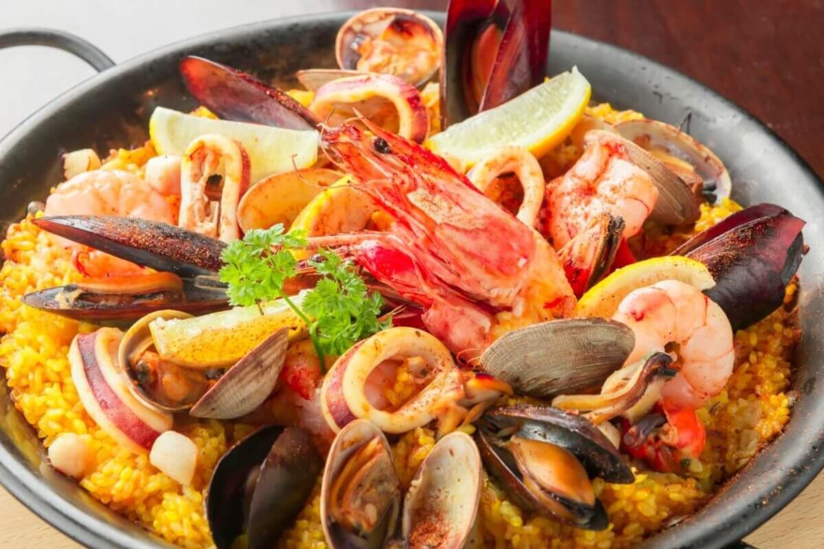 A black skillet filled with vibrant Spanish seafood paella showcases a sulit dining experience. The dish features yellow saffron rice topped generously with mussels, clams, shrimp, squid, and slices of lemon. A sprig of parsley garnishes the center. Ideal for those exploring the best restaurants in Muntinlupa City.