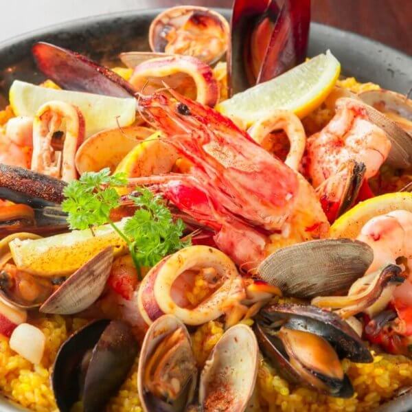 A black skillet filled with vibrant Spanish seafood paella showcases a sulit dining experience. The dish features yellow saffron rice topped generously with mussels, clams, shrimp, squid, and slices of lemon. A sprig of parsley garnishes the center. Ideal for those exploring the best restaurants in Muntinlupa City.