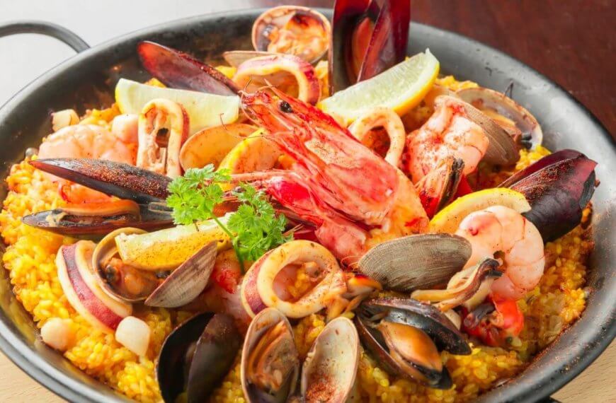 A black skillet filled with vibrant Spanish seafood paella showcases a sulit dining experience. The dish features yellow saffron rice topped generously with mussels, clams, shrimp, squid, and slices of lemon. A sprig of parsley garnishes the center. Ideal for those exploring the best restaurants in Muntinlupa City.