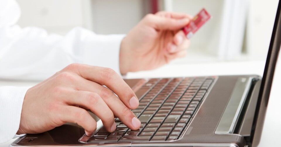 A person in a white shirt is using a laptop, typing with one hand while holding a red credit card in the other. In an indoor setting atop a white surface, this scenario offers practical tips for small business owners looking to upgrade their online stores. The focus remains on the hands and the keyboard.