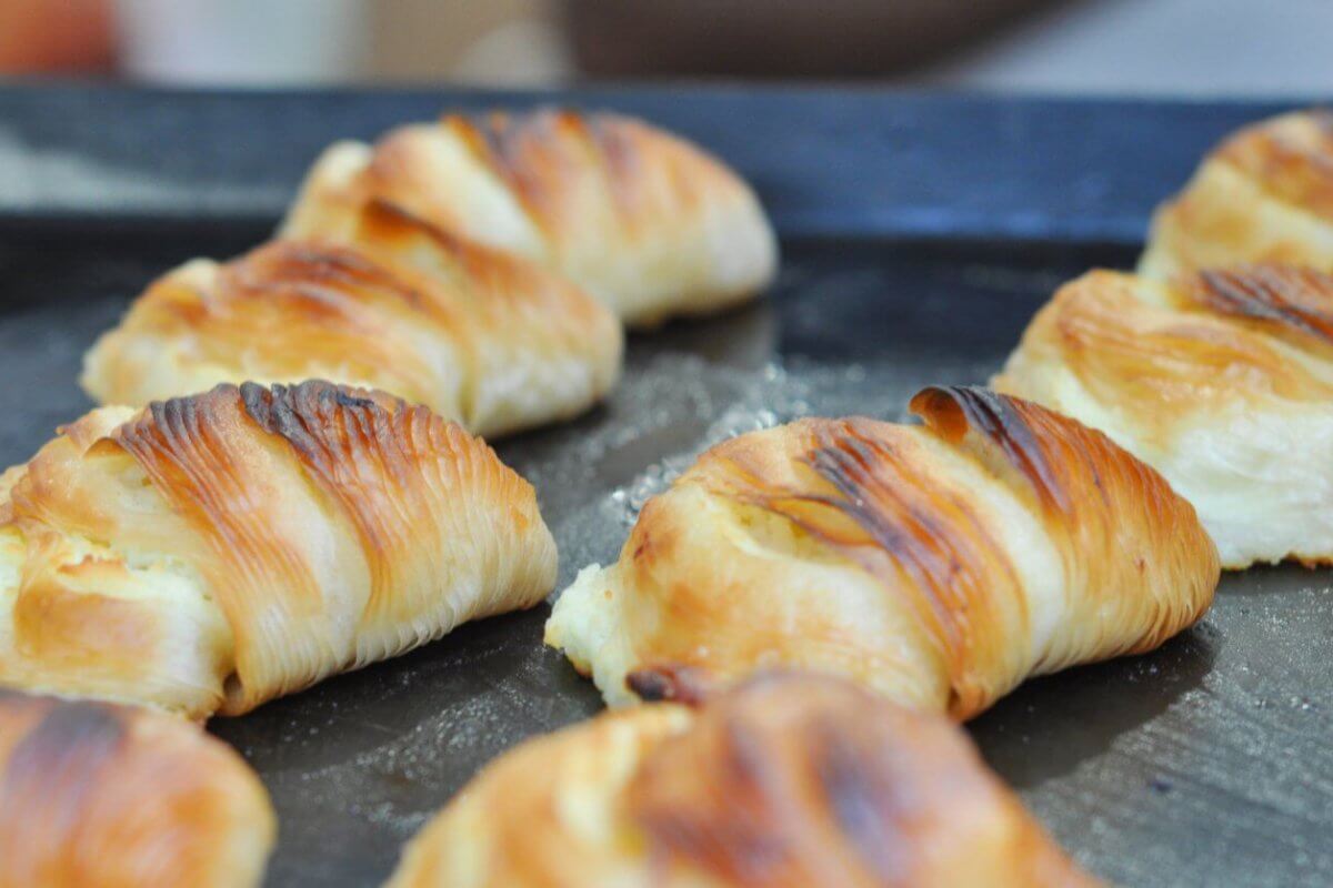 A close-up of golden-brown croissants lined up on a dark baking tray. The croissants appear freshly baked with flaky, buttery layers and slightly crispy, browned tops. Perfect for those sweet cravings, these treats are sourced from top pastry suppliers in Metro Manila.
