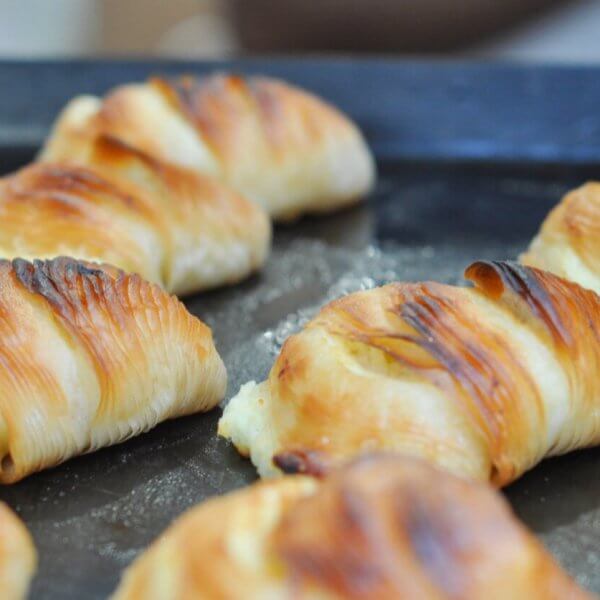 A close-up of golden-brown croissants lined up on a dark baking tray. The croissants appear freshly baked with flaky, buttery layers and slightly crispy, browned tops. Perfect for those sweet cravings, these treats are sourced from top pastry suppliers in Metro Manila.