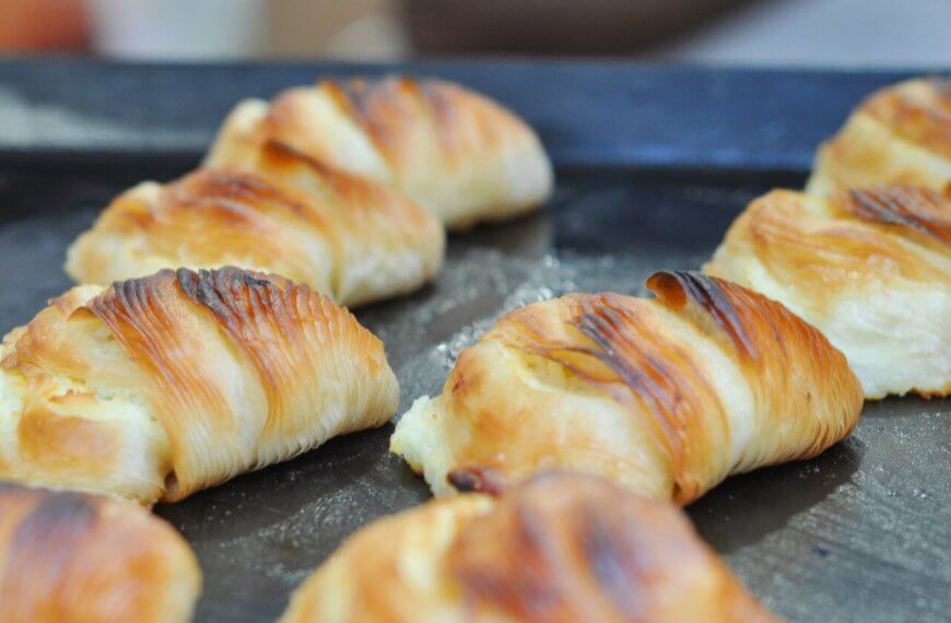A close-up of golden-brown croissants lined up on a dark baking tray. The croissants appear freshly baked with flaky, buttery layers and slightly crispy, browned tops. Perfect for those sweet cravings, these treats are sourced from top pastry suppliers in Metro Manila.