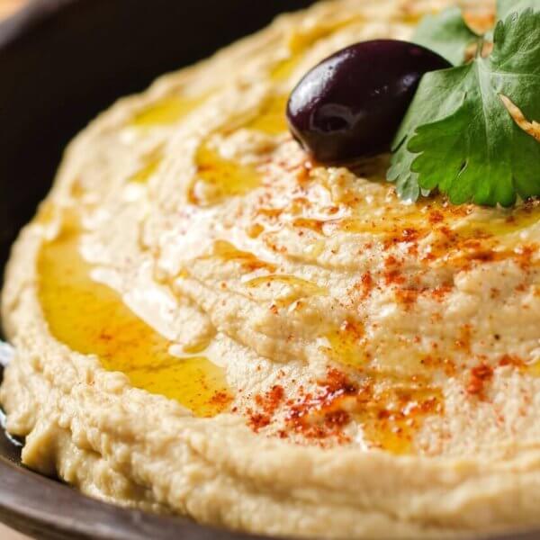A close-up shot of a bowl of creamy hummus, garnished with two black olives, a sprig of fresh parsley, and a drizzle of olive oil. A sprinkle of paprika adds a touch of color on top. The dish is presented in a dark bowl, with blurred crackers visible in the background—a true culinary adventure.