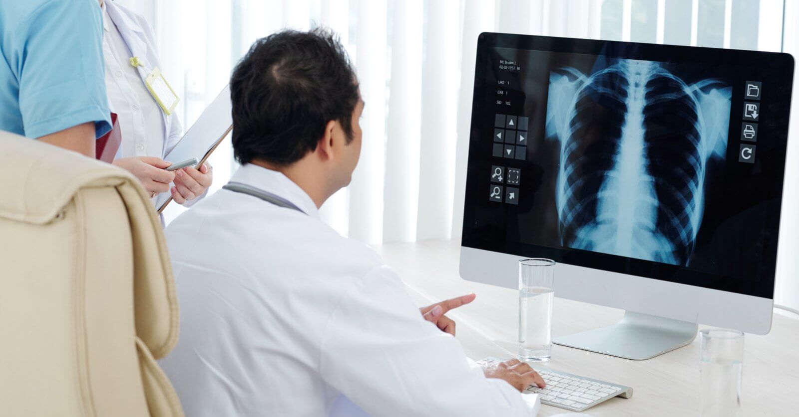 A doctor in a white coat, recognized as the best pulmonologist in Metro Manila, examines a chest X-ray image displayed on a computer monitor. Next to the doctor, a nurse in a blue uniform holds a clipboard and stands. A glass of water and keyboard are on the desk. Vertical blinds cover the window in the background.