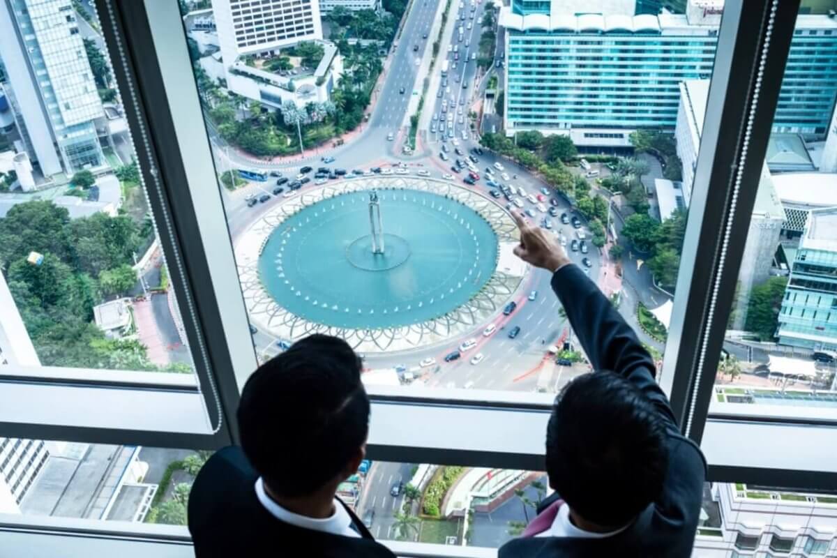 Two individuals in business attire are standing beside large windows in a high-rise building, overlooking the bustling cityscape of Metro Manila. One person is pointing toward a circular fountain surrounded by traffic on a busy street below. Tall buildings, greenery, and various vehicles are visible, highlighting the prime locations often sought by realty companies.