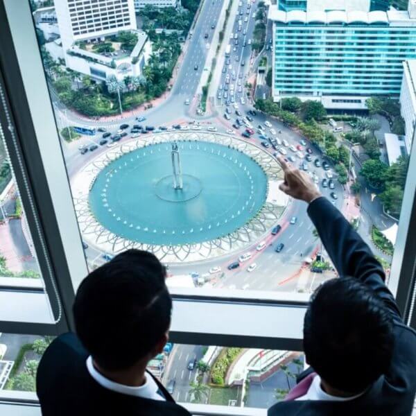 Two individuals in business attire are standing beside large windows in a high-rise building, overlooking the bustling cityscape of Metro Manila. One person is pointing toward a circular fountain surrounded by traffic on a busy street below. Tall buildings, greenery, and various vehicles are visible, highlighting the prime locations often sought by realty companies.