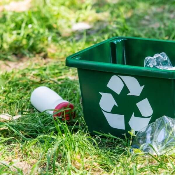 A green recycling bin with a white recycling symbol lies on the grass outdoors in Metro Manila. The bin contains a crumpled plastic bottle and is surrounded by scattered litter, including another crumpled plastic bottle and a white bottle with a red cap. Sunlight filters through the grassy area.
