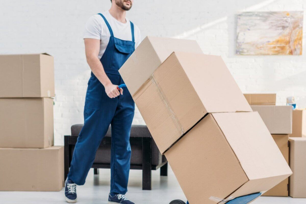 A person wearing a white t-shirt and blue overalls pushes a hand truck loaded with three large cardboard boxes. Several more boxes are stacked in the background of the room decorated with white walls and a modern abstract painting, indicating the provided relocation services. The person appears to be engaged in moving or packing activities.