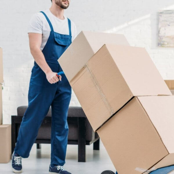 A person wearing a white t-shirt and blue overalls pushes a hand truck loaded with three large cardboard boxes. Several more boxes are stacked in the background of the room decorated with white walls and a modern abstract painting, indicating the provided relocation services. The person appears to be engaged in moving or packing activities.