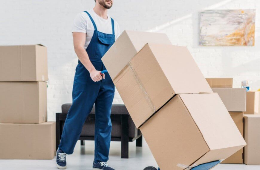 A person wearing a white t-shirt and blue overalls pushes a hand truck loaded with three large cardboard boxes. Several more boxes are stacked in the background of the room decorated with white walls and a modern abstract painting, indicating the provided relocation services. The person appears to be engaged in moving or packing activities.