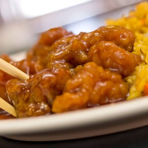 A plate featuring a serving of sweet and sour chicken, with pieces glistening in sauce and being picked up with a pair of chopsticks. On the side is a portion of fried rice, colorful with bits of vegetables like carrots and peas. This dish could be found at one of the best restaurants near Manila's 168 Shopping Mall.