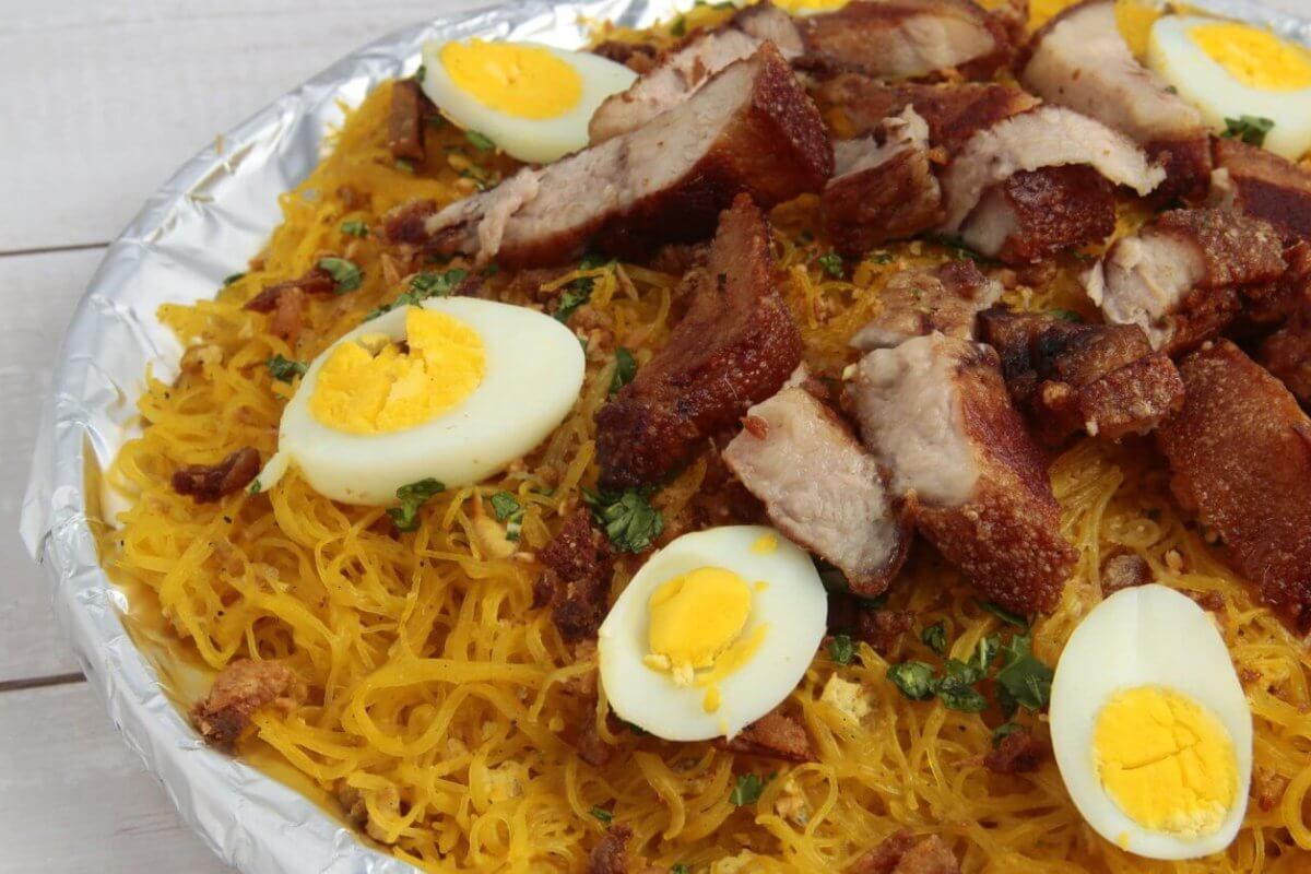 A plate filled with a Filipino noodle dish called Pancit Malabon, featuring yellow rice noodles topped with sliced hard-boiled eggs, crispy pork belly, cooked shrimp, and chopped green onions. The elements are arranged aesthetically on a white wooden table near the popular restos along Taft Avenue.