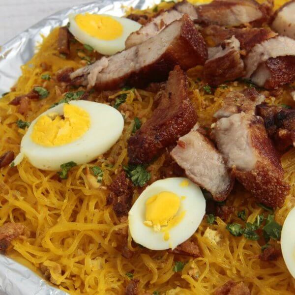 A plate filled with a Filipino noodle dish called Pancit Malabon, featuring yellow rice noodles topped with sliced hard-boiled eggs, crispy pork belly, cooked shrimp, and chopped green onions. The elements are arranged aesthetically on a white wooden table near the popular restos along Taft Avenue.