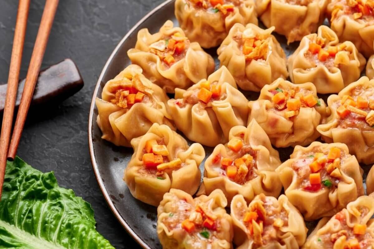 A plate filled with neatly arranged steamed dumplings, each topped with a small garnish of finely chopped vegetables. Wooden chopsticks rest on a holder beside the plate. A fresh green lettuce leaf is placed adjacent, providing a touch of color to the dark background at New Greenhills Mall's 84 restaurants.