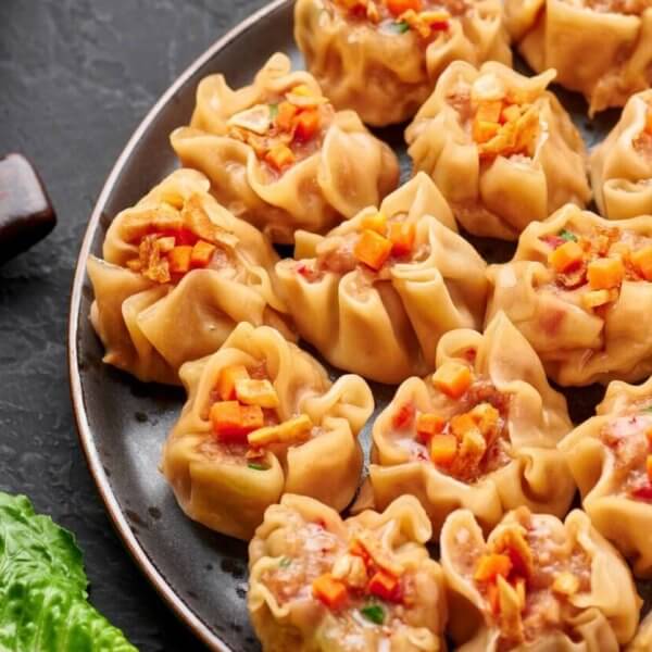 A plate filled with neatly arranged steamed dumplings, each topped with a small garnish of finely chopped vegetables. Wooden chopsticks rest on a holder beside the plate. A fresh green lettuce leaf is placed adjacent, providing a touch of color to the dark background at New Greenhills Mall's 84 restaurants.