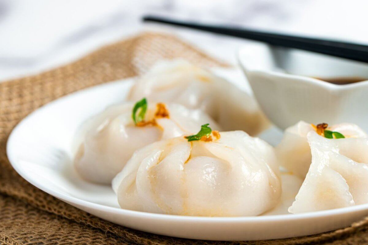 Hakaw steamed dumplings garnished with small green herbs, accompanies by a dipping sauce in a white bowl, and a pair of black chopsticks resting across it. The setup is styled on burlap fabric atop a marble surface—an elegant touch reminiscent of Top Restaurants found in Power Plant Mall.