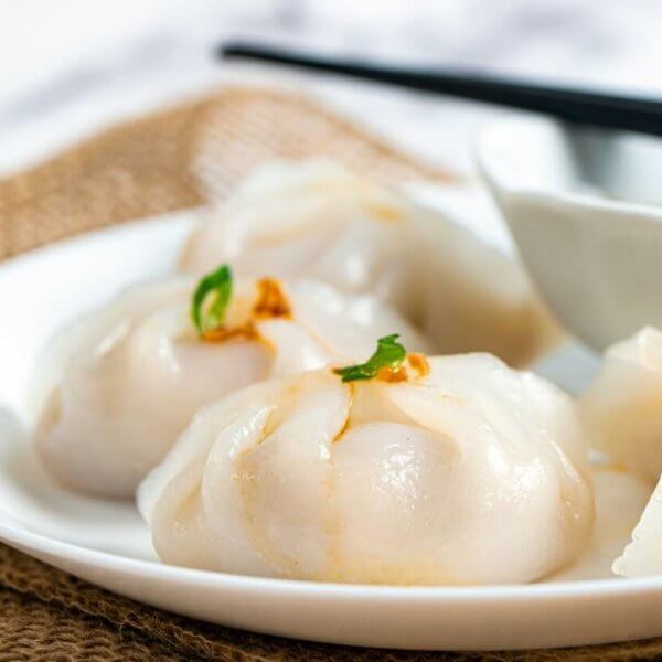 Hakaw steamed dumplings garnished with small green herbs, accompanies by a dipping sauce in a white bowl, and a pair of black chopsticks resting across it. The setup is styled on burlap fabric atop a marble surface—an elegant touch reminiscent of Top Restaurants found in Power Plant Mall.