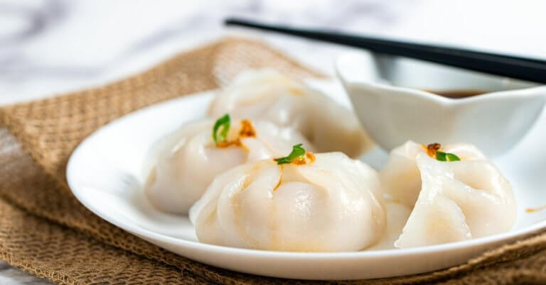 Hakaw steamed dumplings garnished with small green herbs, accompanies by a dipping sauce in a white bowl, and a pair of black chopsticks resting across it. The setup is styled on burlap fabric atop a marble surface—an elegant touch reminiscent of Top Restaurants found in Power Plant Mall.