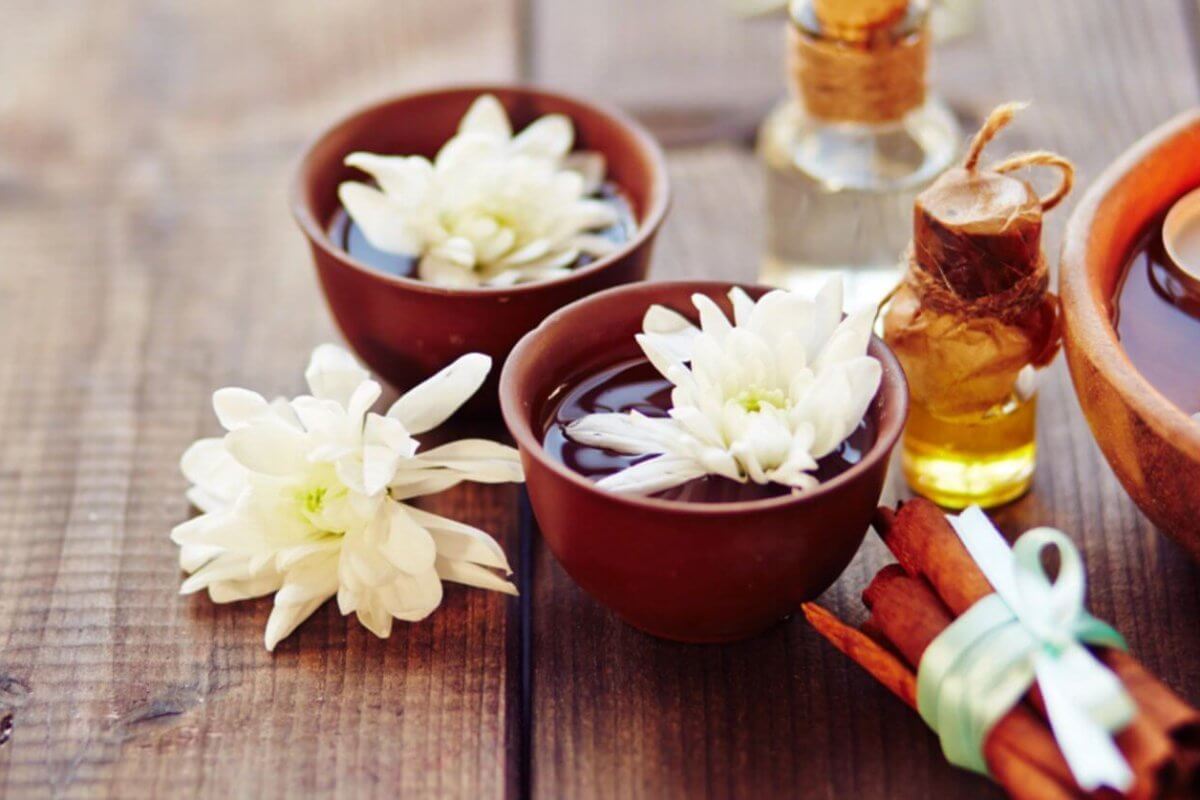 A serene spa scene on a wooden surface features two brown bowls filled with water and white daisies, a lit candle in another bowl, and a small glass bottle with a cork stopper containing oil. A bundle of cinnamon sticks tied with a green ribbon lies nearby, adding to the tranquil setup sourced from the finest fragrance suppliers in Metro Manila.