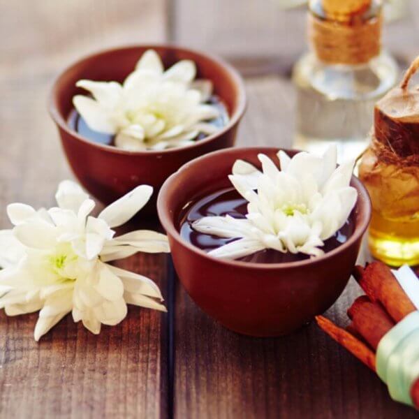 A serene spa scene on a wooden surface features two brown bowls filled with water and white daisies, a lit candle in another bowl, and a small glass bottle with a cork stopper containing oil. A bundle of cinnamon sticks tied with a green ribbon lies nearby, adding to the tranquil setup sourced from the finest fragrance suppliers in Metro Manila.