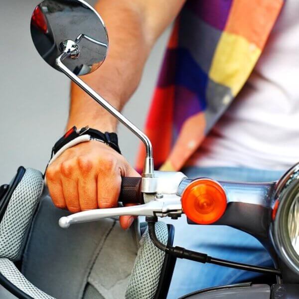 Close-up of a person wearing a colorful plaid shirt and a white T-shirt holding the handlebar of a scooter from one of the top scooter shops in Metro Manila. The focus is on their left hand gripping the handle while wearing a black wristwatch. The scooter features a large headlight, an orange turn signal, and a rearview mirror.