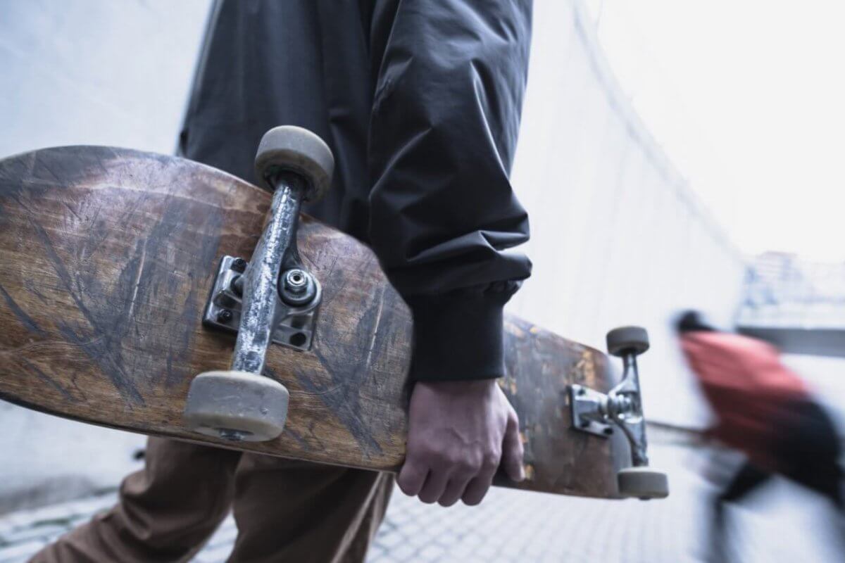 A person in a dark jacket holds a worn skateboard by their side while walking on a paved pathway. The background is a blurred image of another person in motion, wearing a red jacket, moving down a sloped area. The urban setting with concrete structures could be reminiscent of Metro Manila's bustling vibe.