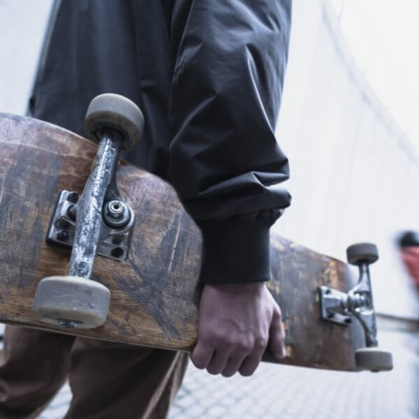 A person in a dark jacket holds a worn skateboard by their side while walking on a paved pathway. The background is a blurred image of another person in motion, wearing a red jacket, moving down a sloped area. The urban setting with concrete structures could be reminiscent of Metro Manila's bustling vibe.