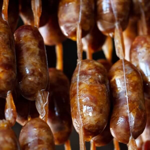Close-up of several rows of sausages hanging vertically by beige strings. The sausages have a brownish color with visible spices embedded in the meat. The links appear glossy and slightly uneven, indicating they are likely homemade or artisanal. This delicacy could be a highlight at one of Davao's 42 Best Restaurants in SM Lanang.
