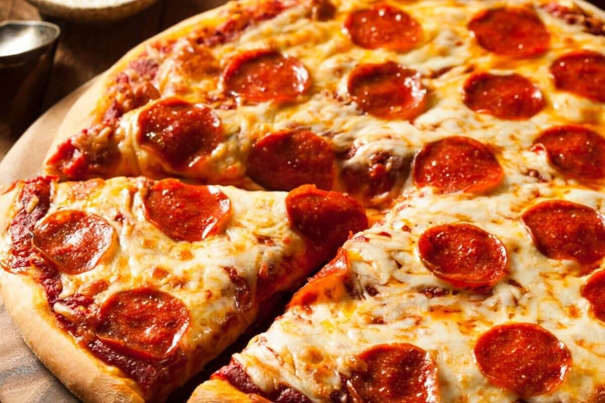 A close-up of a pepperoni pizza on a wooden surface. The pizza has a golden-brown crust and is topped with melted cheese and evenly distributed pepperoni slices. One slice is partially separated from the rest, showcasing the gooey cheese and crisp edges. Leafy greens and utensils are blurred in the background, reminiscent of a Sulit Dining Experience at SM Manila's best restaurants.