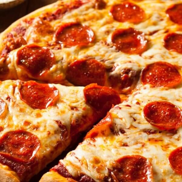 A close-up of a pepperoni pizza on a wooden surface. The pizza has a golden-brown crust and is topped with melted cheese and evenly distributed pepperoni slices. One slice is partially separated from the rest, showcasing the gooey cheese and crisp edges. Leafy greens and utensils are blurred in the background, reminiscent of a Sulit Dining Experience at SM Manila's best restaurants.