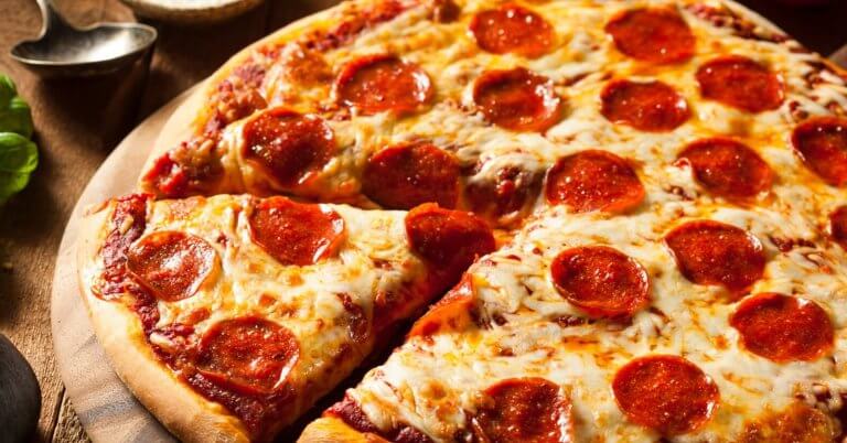 A close-up of a pepperoni pizza on a wooden surface. The pizza has a golden-brown crust and is topped with melted cheese and evenly distributed pepperoni slices. One slice is partially separated from the rest, showcasing the gooey cheese and crisp edges. Leafy greens and utensils are blurred in the background, reminiscent of a Sulit Dining Experience at SM Manila's best restaurants.