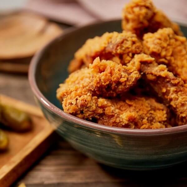 A bowl filled with crispy fried chicken pieces sits on a wooden surface. Beside it, a wooden tray holds several pickles and some fries. In the background, a wooden spoon and a jar of dipping sauce can be seen. This cozy, rustic scene captures the essence of comfort food you'd find at one of the best restaurants in SM Podium Mall.