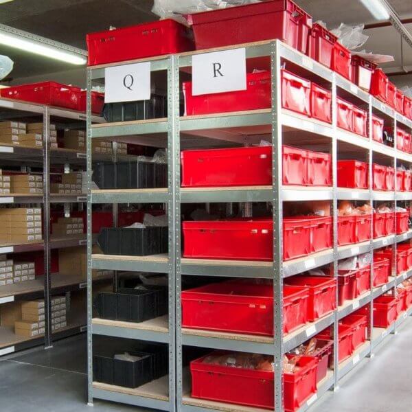 A well-organized warehouse aisle in Metro Manila features metal shelving units filled with red and black plastic storage bins. Each shelf has white labels with letters "Q" and "R" indicating storage categories. Companies rely on these efficient storage solutions, where cardboard boxes and items are neatly arranged in the bins. The floor is clean and polished.