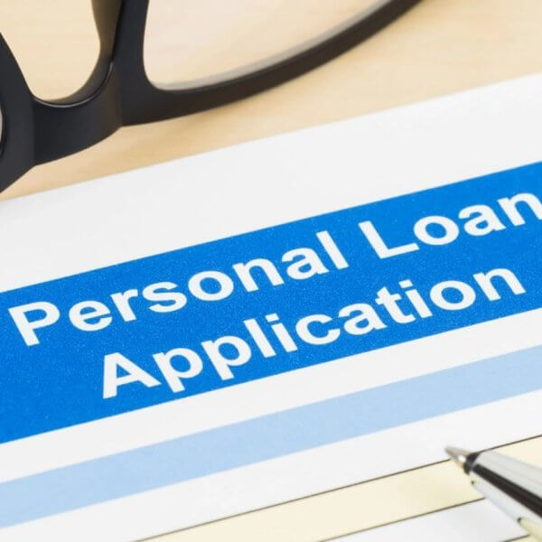 A close-up of a Sulit personal loan application form with a blue header. "Personal Loan Application" is prominently displayed. There's a pen on the form, and a pair of glasses is partially visible in the upper left corner. The form includes fields to fill in personal details alongside the convenient use of a loan calculator.