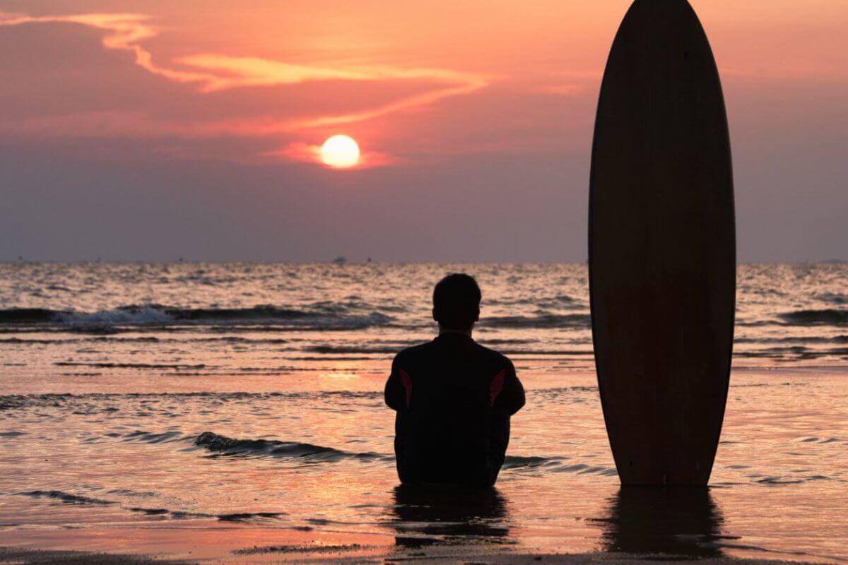 A person, seen from behind, sits on wet sand at the beach during sunset. Next to them, a surfboard is positioned upright in the sand. The sky is filled with vibrant hues of orange and pink as the sun sets over the calm ocean waves in the background, evoking memories of days at surfing school.