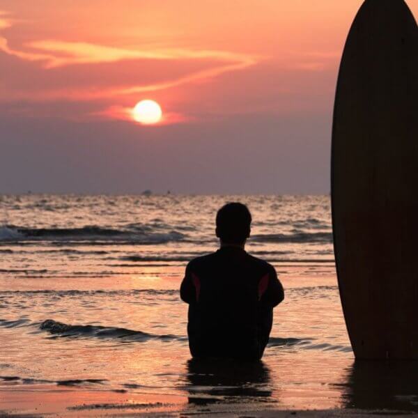 A person, seen from behind, sits on wet sand at the beach during sunset. Next to them, a surfboard is positioned upright in the sand. The sky is filled with vibrant hues of orange and pink as the sun sets over the calm ocean waves in the background, evoking memories of days at surfing school.