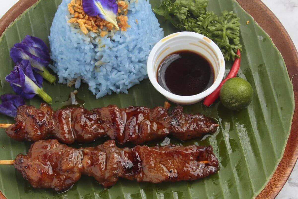 A plate with two bbq and blue rice garnished with crispy bits and an edible flower, a small cup of dark dipping sauce, a lime, a chili, and parsley served on a large banana leaf. Celebrated among the 50 Best Restaurants near UP Diliman, this dish is presented on a wooden tray against a light background.