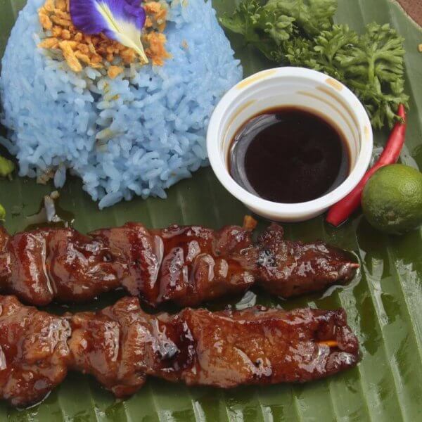 A plate with two bbq and blue rice garnished with crispy bits and an edible flower, a small cup of dark dipping sauce, a lime, a chili, and parsley served on a large banana leaf. Celebrated among the 50 Best Restaurants near UP Diliman, this dish is presented on a wooden tray against a light background.
