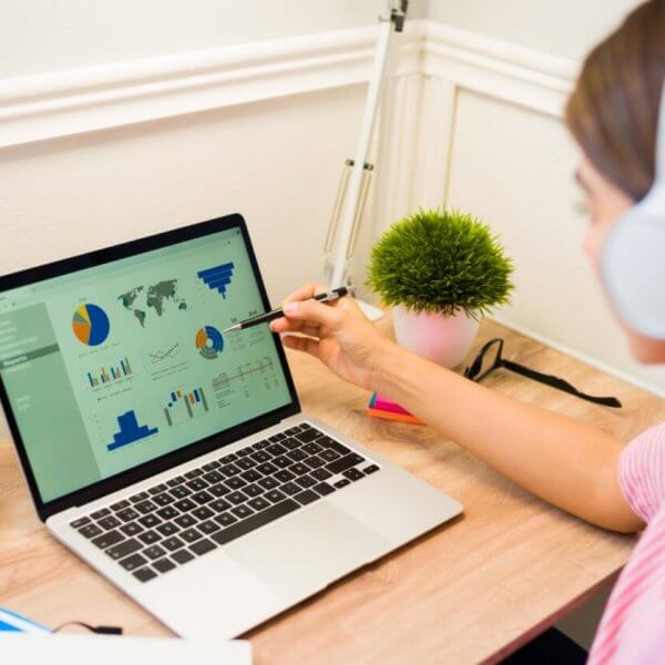 Virtual Assistant wearing headphones sits at a desk in Cebu, focusing on a laptop displaying various graphs and charts. They are pointing to the screen with a pen. The workspace includes colorful sticky notes, a pot of pens, and a small potted plant. The room is well-lit from a nearby window, perfect for productivity with the best virtual assistants.